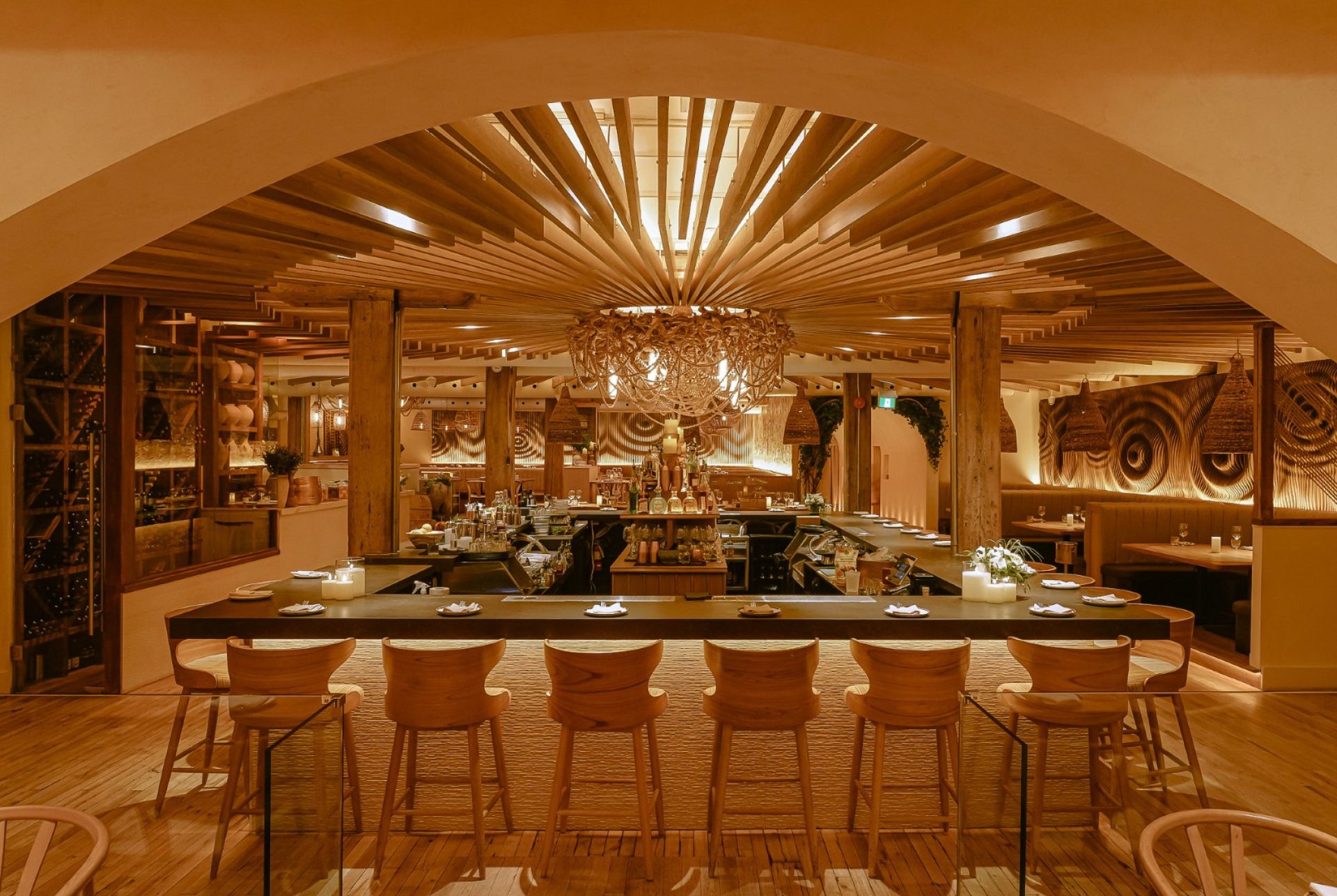 A warmly lit restaurant interior with a long central bar and wood stools facing an open kitchen. Curved arches, layered wood ceiling details, and decorative lighting create an intimate, upscale dining atmosphere.